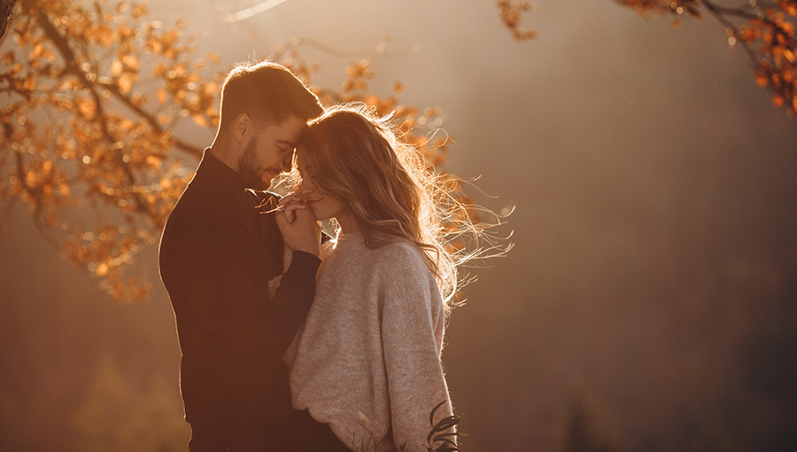 Stylish young couple in the autumn mountains. A guy and a girl hug together under a large old tree on a background of a forest and mountain peaks at sunset.