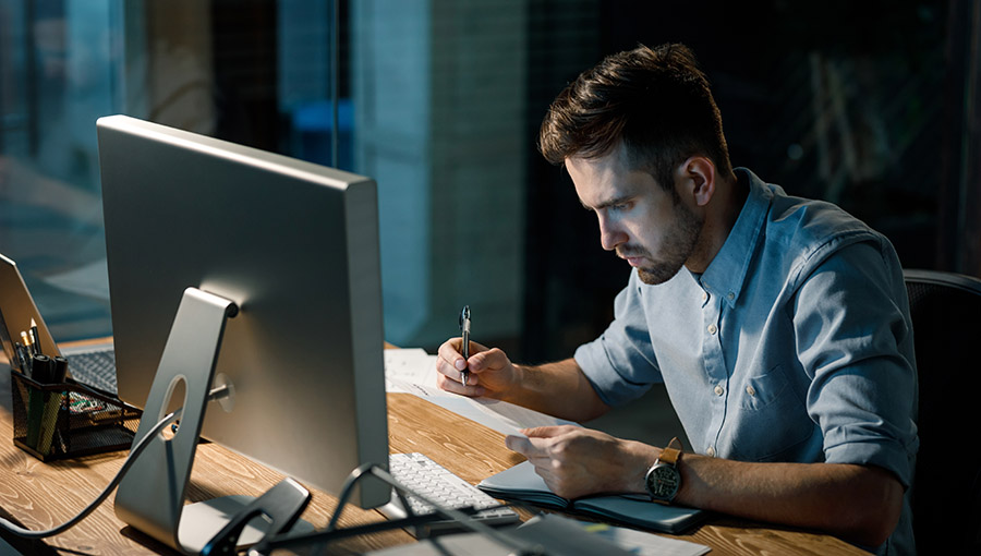 Handsome young man sitting at desk with computer taking notes while working overhours on important project. 