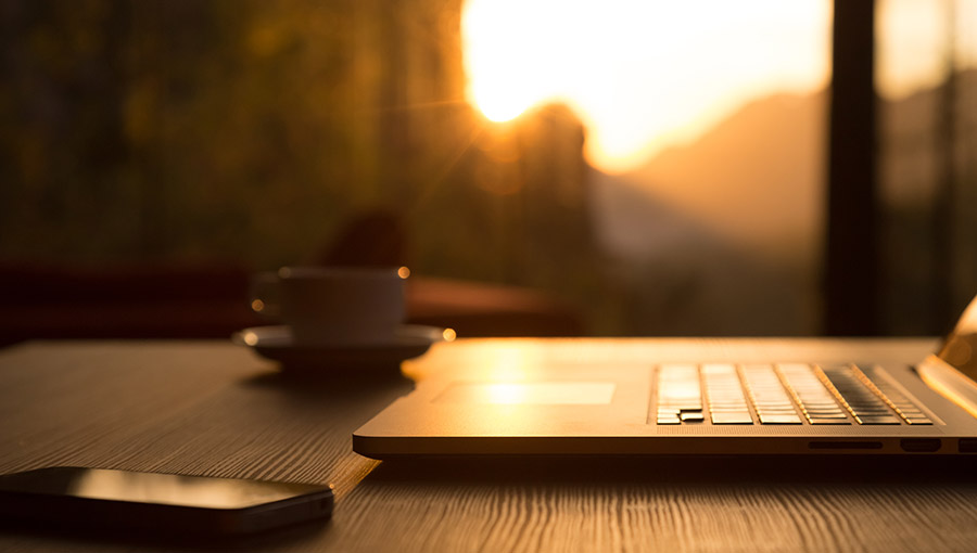 Nomad Work Concept Image Computer Coffee Mug and Telephone on black wood Table and Evening Sunlight shining throw large windows focus on Edge of Laptop